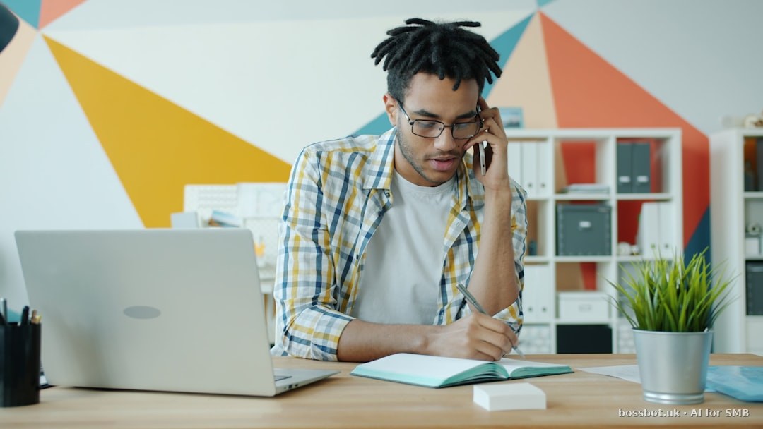 Man talking on phone at desk with laptop and notebook.